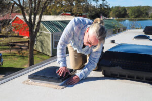 Person inspecting a roof vent on top of an RV parked near a lake and campground.