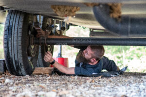 Technician lying on the ground beneath an RV inspecting or repairing something near the rear wheels.