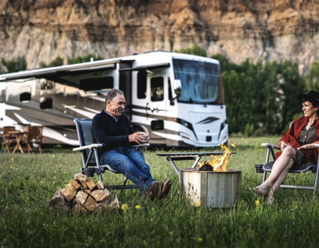 Two people sitting in a green field around a fire in front of an RV.