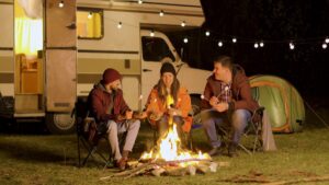 Go RVing’s Trends To Know: Community 2 Group of happy friends around burning camping fire in the woods roasting marshmallows. Retro camper van.