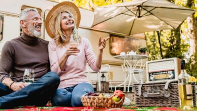 Aged beautiful caucasian couple having picnic and drinking champagne in the yard near the motorhome.Mature blond lady pointing at something