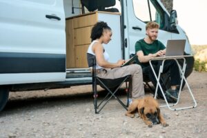 Young couple working remotely using laptop and tablet near their camper van with their dog resting nearby.