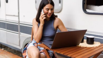 Young woman working on laptop talking on phone near her motorhome to illustrate a digital nomad lifestyle.