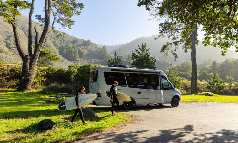 A Leisure Travel Vans RV in the wilderness, with two people carrying surfboards walking away from the RV.