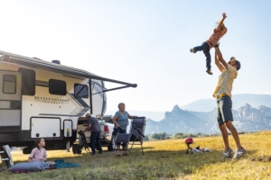 RV in background with mom and kid sitting outside RV in camp chairs and dad standing up holding child above his head with mountains in background.
